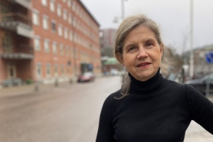 Portrait of Helén Sjöland wearing a black turtleneck and shot from the torso up, outdoors with a street and building in the background.